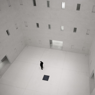 Uma sala branca com duas portas e diversas janelas com uma única pessoa de preto no centro